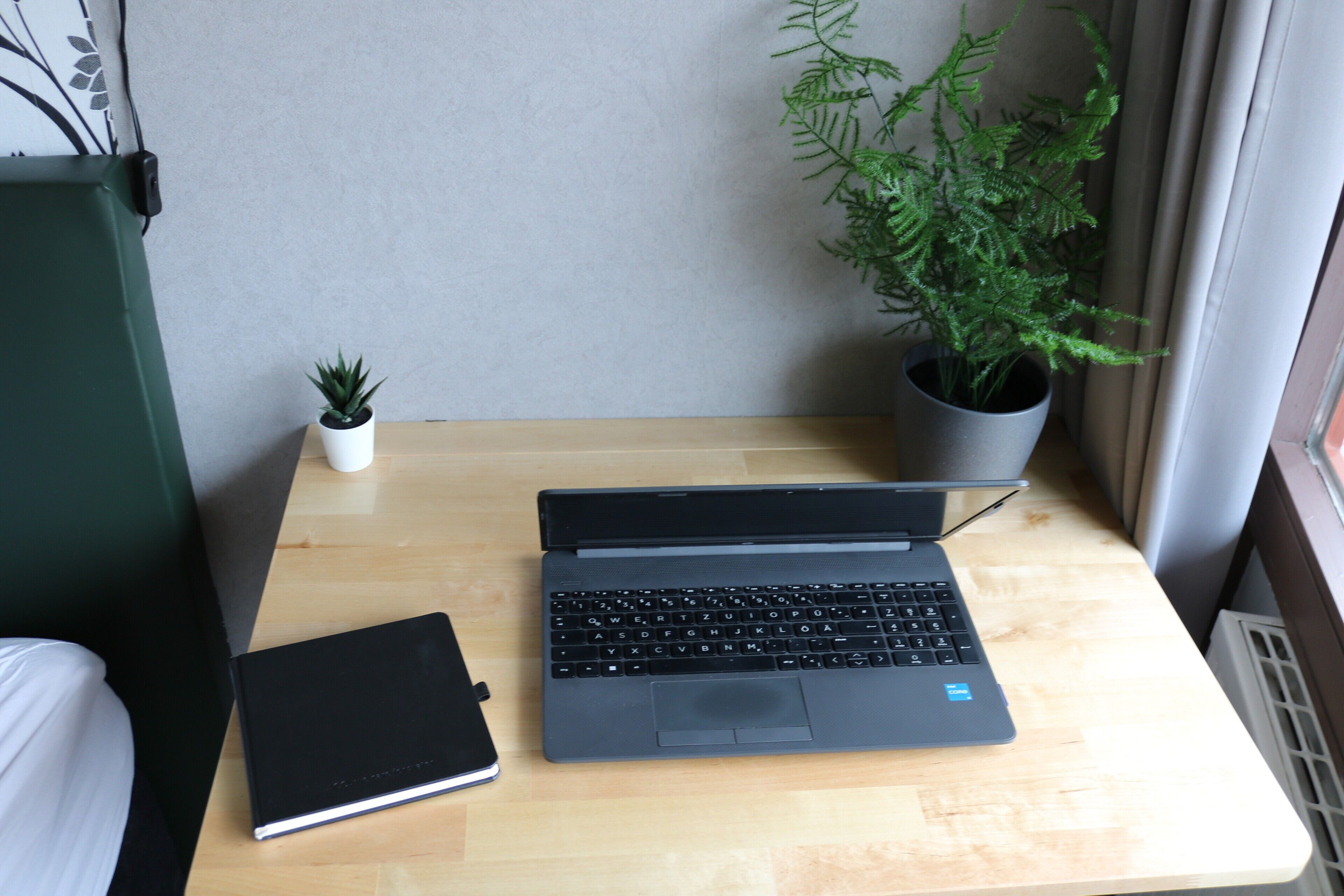 A laptop on a wooden desk with a potted plant and a notebook beside it.