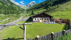 Una cabaña con fachada de madera, un patio con mobiliario exterior y montañas al fondo.