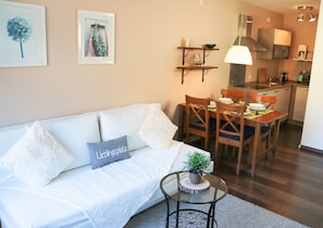 A modern living room with a white sofa, a dining table with chairs, a small side table with a plant, and a kitchen area in the background. - Apartment Caroline with Pool & Sauna (Schluchsee)