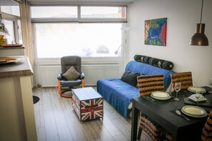 En stue med en sofa, et spisebord dækket til to personer, en radiator, et vindue med persienner og et billede på væggen.