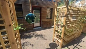 Une pergola en bois surmontée d’un objet sphérique suspendu, une clôture en bois et une porte donnant accès à un bâtiment dont le revêtement extérieur est constitué de panneaux horizontaux.