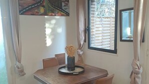 Un coin repas avec une table et des chaises en bois, un vase contenant des fleurs séchées et une fenêtre avec des stores.