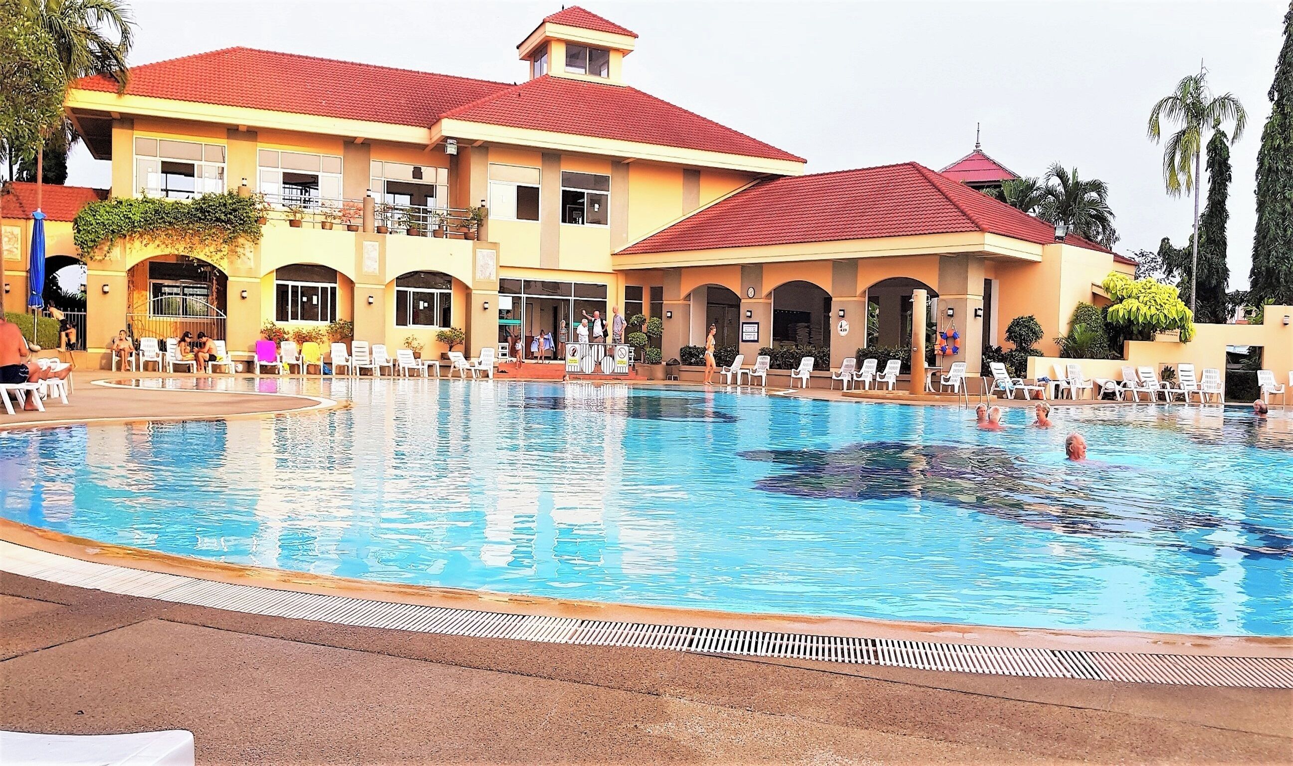 A large outdoor swimming pool with lounge chairs and a building with red-tiled roofs in the background.