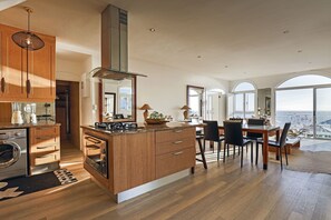 A modern kitchen with wooden cabinets, a central island, and a dining area with a view. - Clifton Sea View Duo Penthouse (Cape Town)