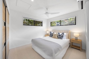A bedroom with a large bed, two bedside tables with lamps, a ceiling fan, and two windows with views of greenery. - Bright Beachside Family House with Ocean Views (Coolum Beach)