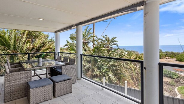 Ein überdachter Balkon mit Korbmöbeln, einem Glastisch und Blick auf Palmen und Meer.