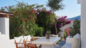 A patio with a white table and chairs, surrounded by lush greenery and pink flowers. - Artemis | 1 Bedroom Apartment with partial mountaView and Patio, Cleopatra Home (Paros)