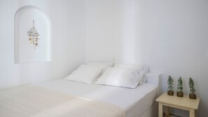 A minimalist bedroom with a bed, white walls, a small bedside table with potted plants, and a wall-mounted light fixture. - Artemis | 1 Bedroom Apartment with partial mountaView and Patio, Cleopatra Home (Paros)