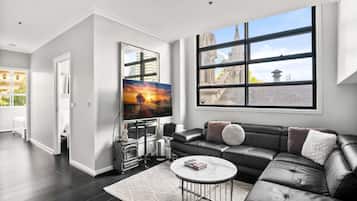 A modern living room with a black leather sofa, a flat-screen TV, and a large window with a view of a building.