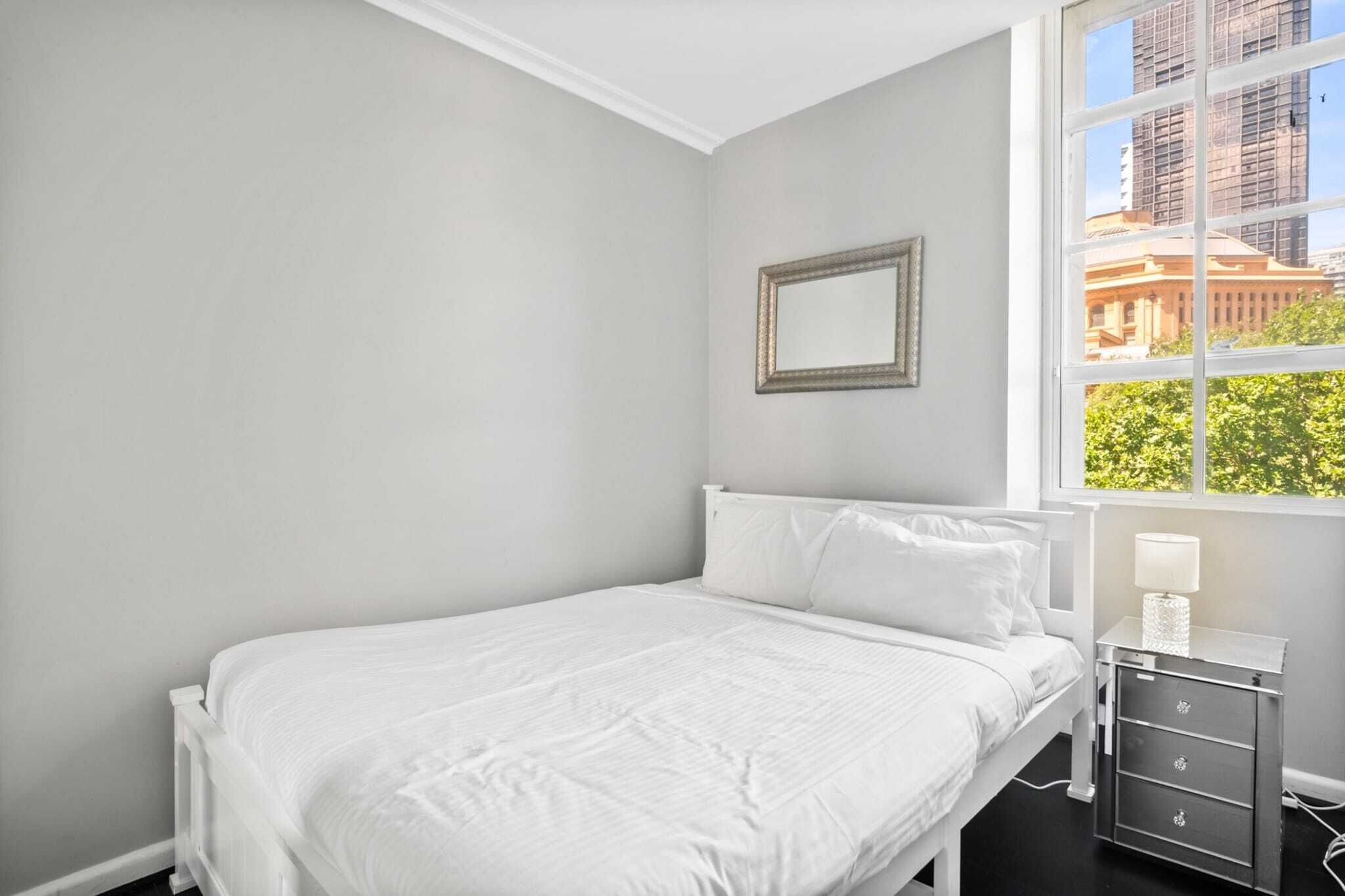 Une chambre à coucher comprenant un lit, une table de chevet, un miroir et une fenêtre donnant sur des immeubles et des arbres.