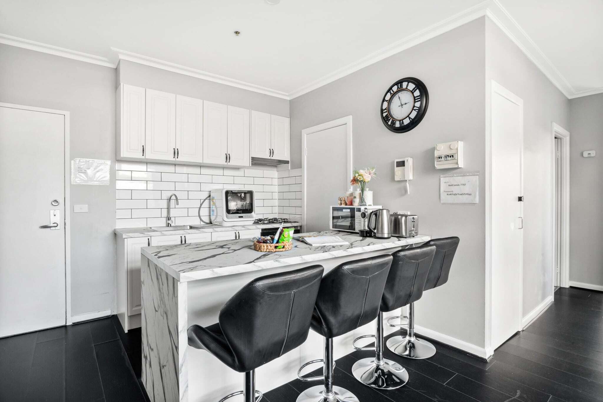 A modern kitchen with white cabinets, a marble island with bar stools, a clock, and a microwave.