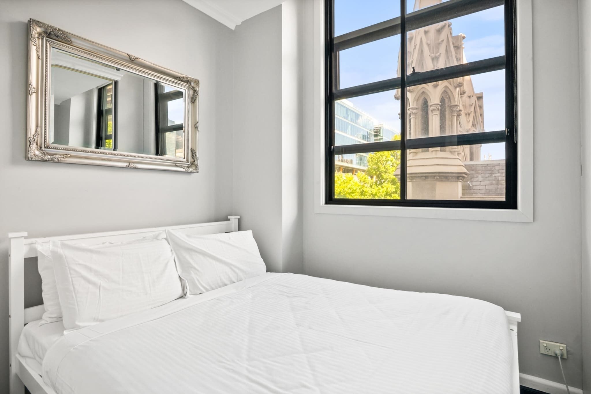 A bedroom with a large window, a bed with white bedding, and a mirror on the wall.