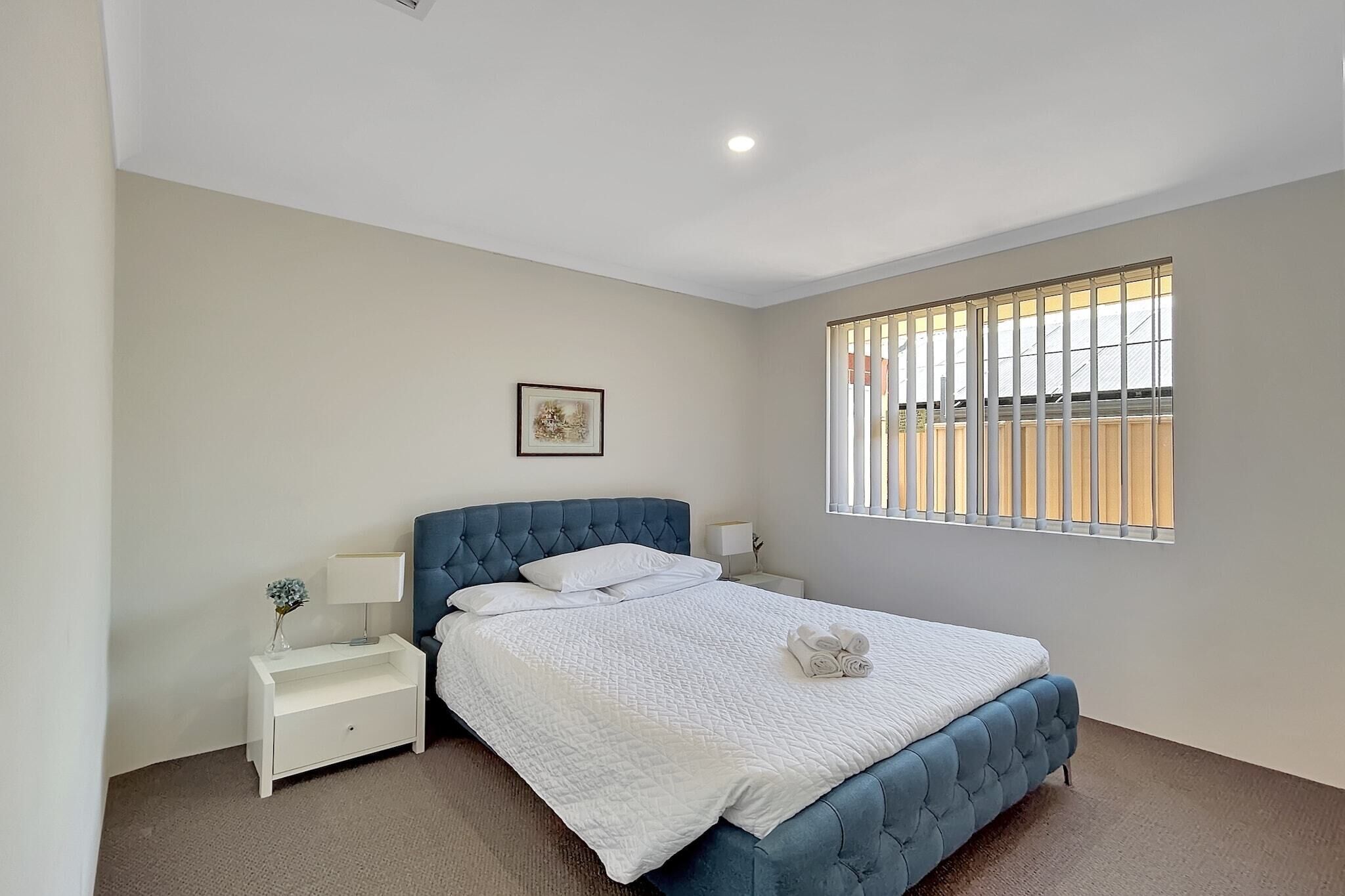 A bedroom with a bed, white nightstands, a picture on the wall, and a window with blinds.