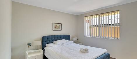 A bedroom with a bed, white nightstands, a picture on the wall, and a window with blinds.