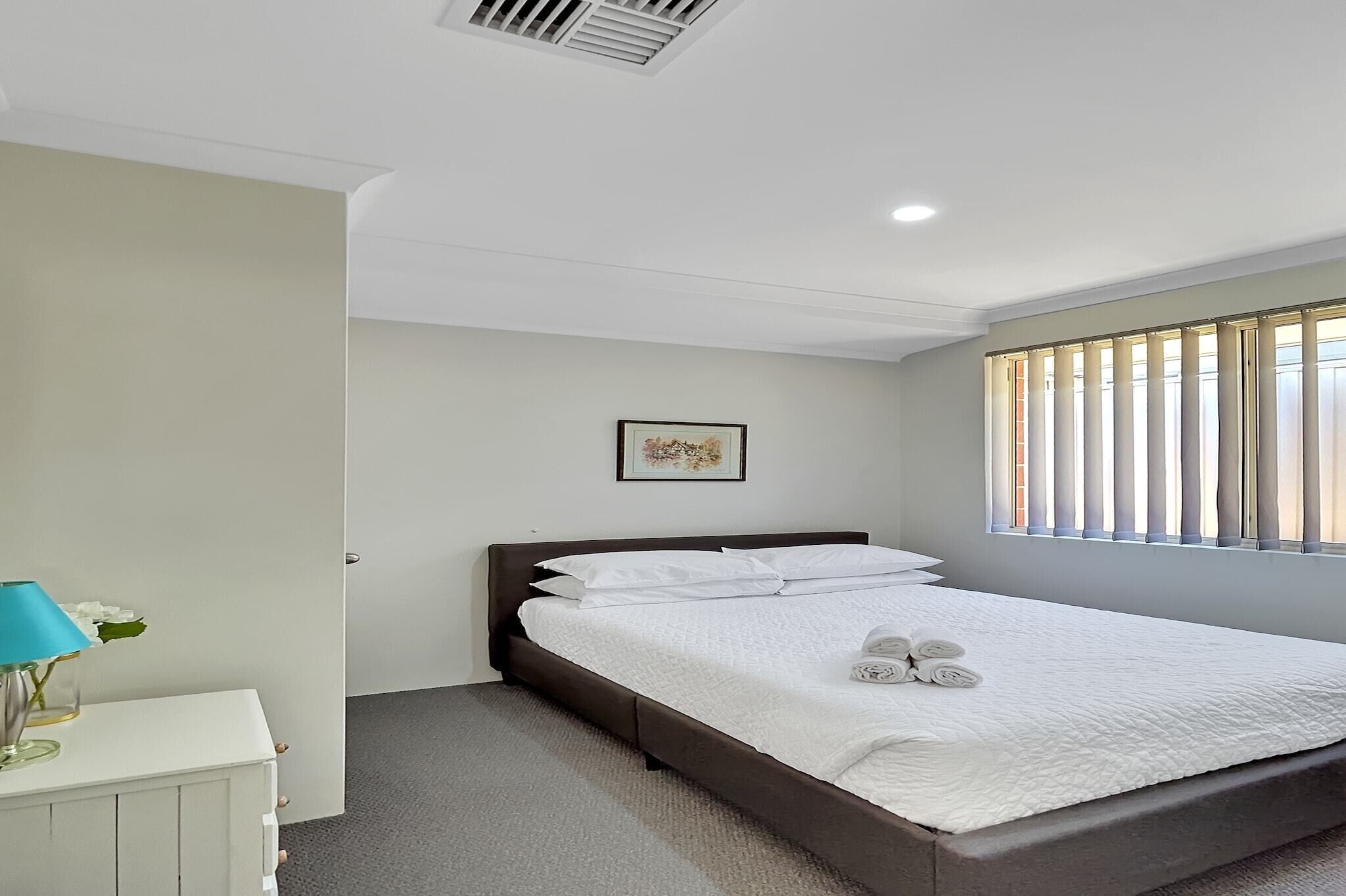 A neatly made bed with white linens and pillows, a bedside table with a lamp and flowers, a framed picture on the wall, and a window with blinds.