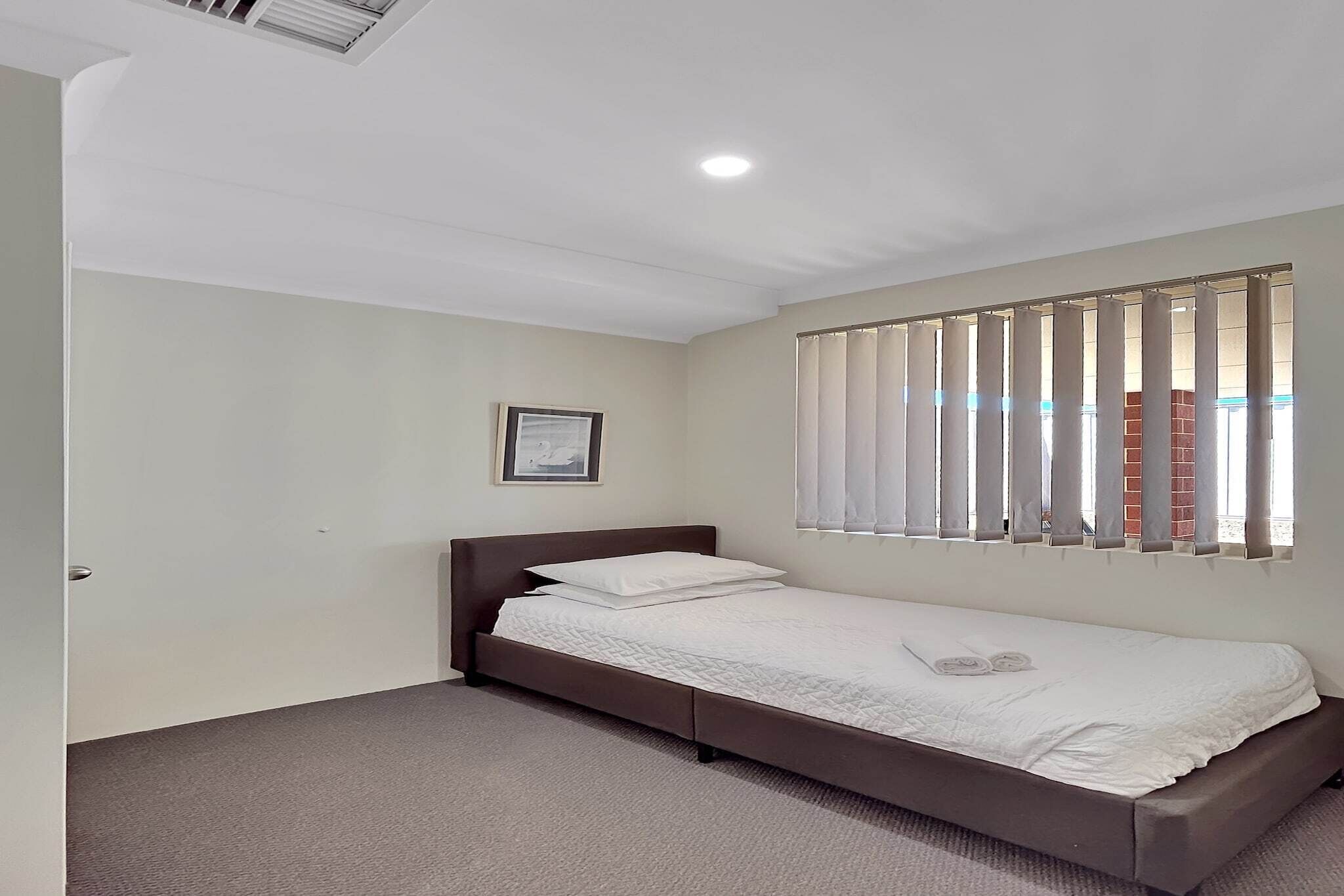 A single bed with a white mattress and pillows, a framed picture on the wall, and a window with vertical blinds.