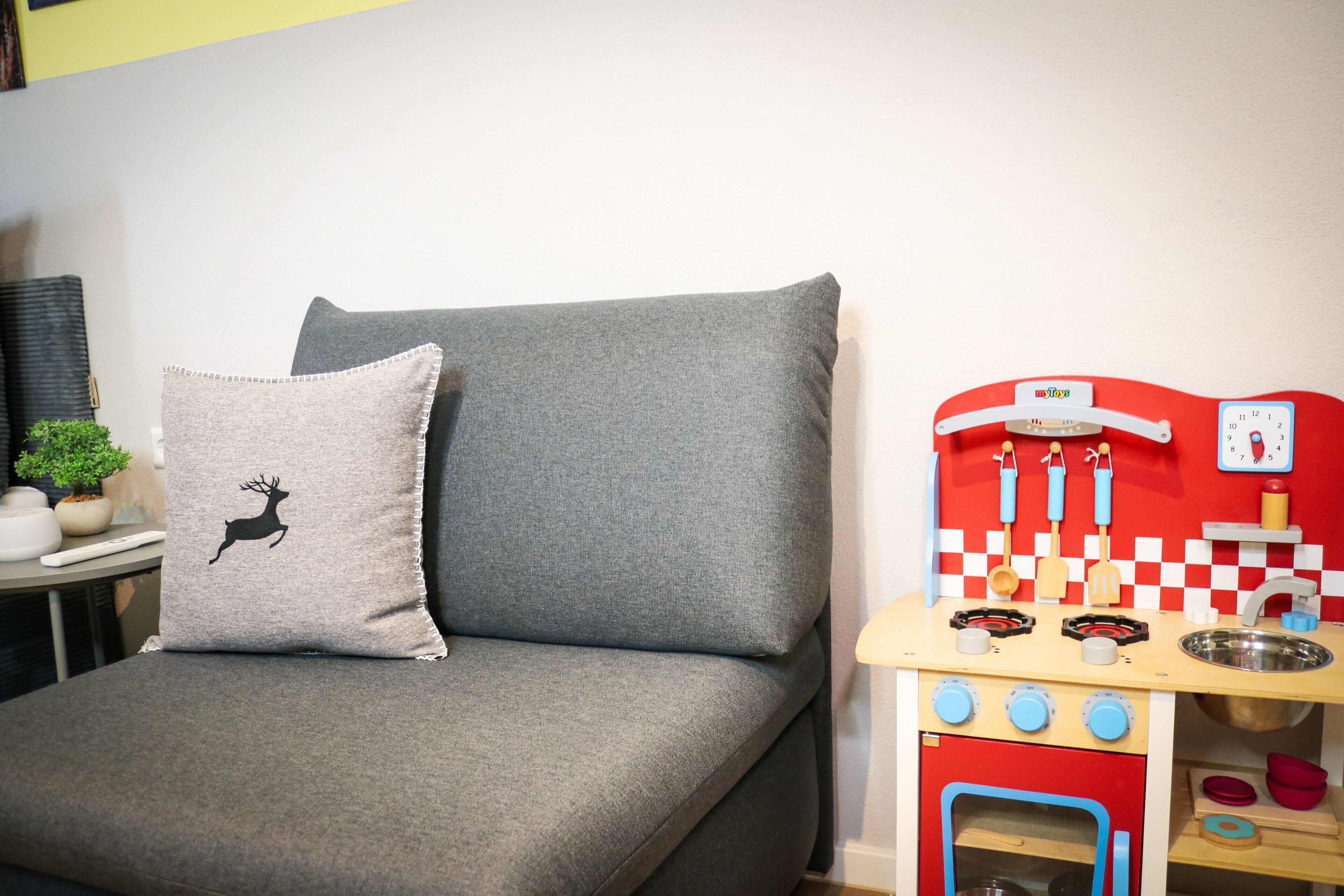 A grey sofa with a pillow featuring a deer silhouette, a small plant, and a toy kitchen set against a white wall.