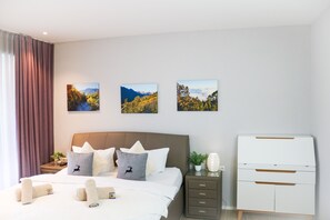 Une chambre d’hôtel moderne, équipée d’un lit, de deux tables de chevet, d’une commode et de trois tableaux représentant des paysages, accrochés au mur.