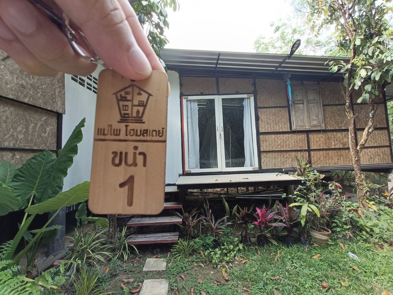 A hand holding a wooden sign with a house icon and text, in front of a house with a window and steps leading up to the door.
