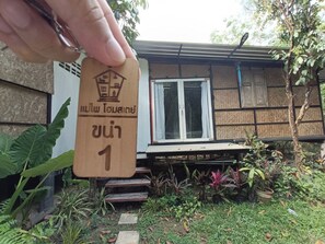 Una mano sostiene un letrero de madera con un icono de casa y texto, frente a una casa con una ventana y escalones que conducen a la puerta.