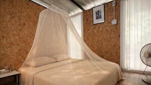 A bedroom with a bed under a mosquito net, wooden walls, a bedside table, a fan, and a framed picture on the wall.