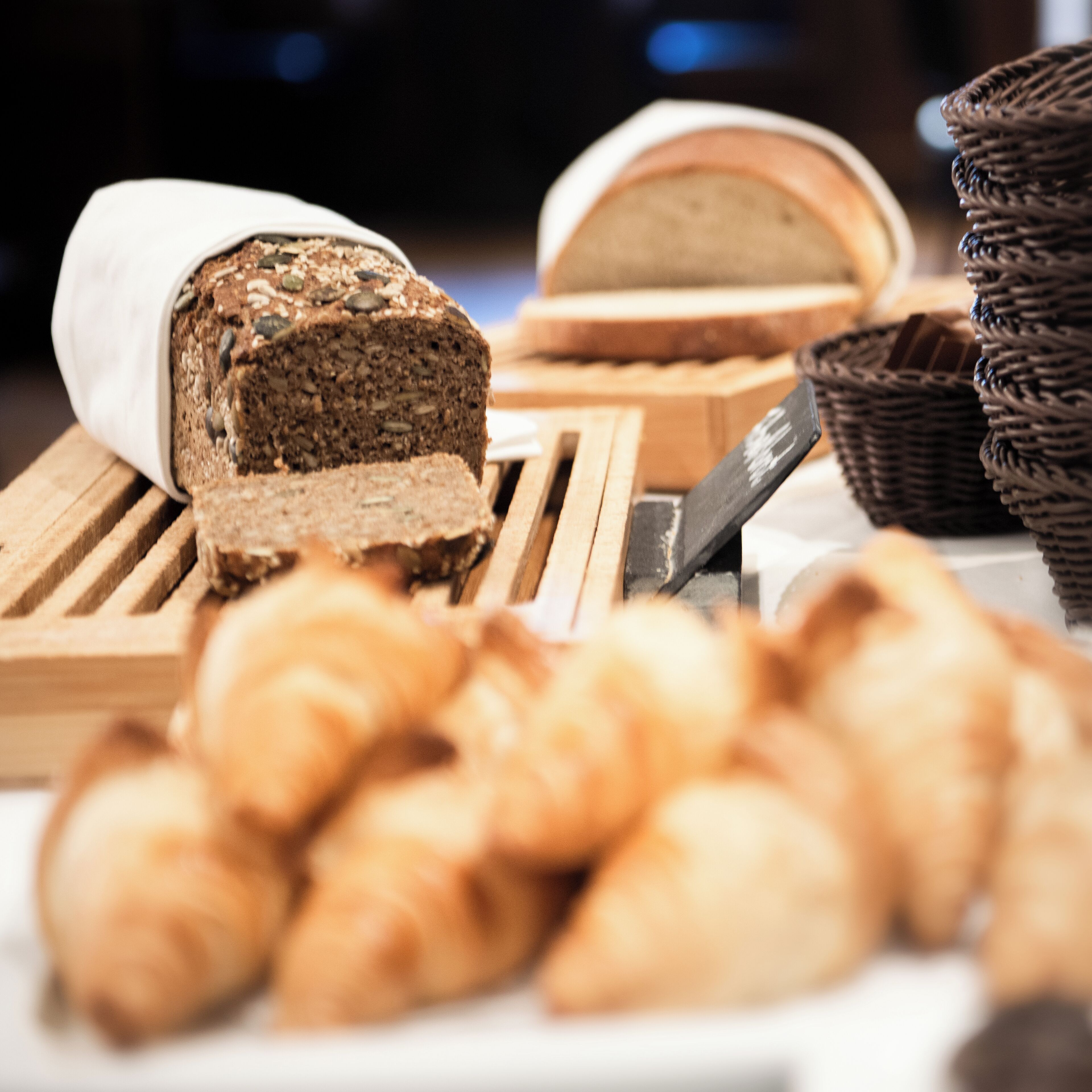 A loaf of bread with seeds on a wooden board, a knife, and several baskets of bread.