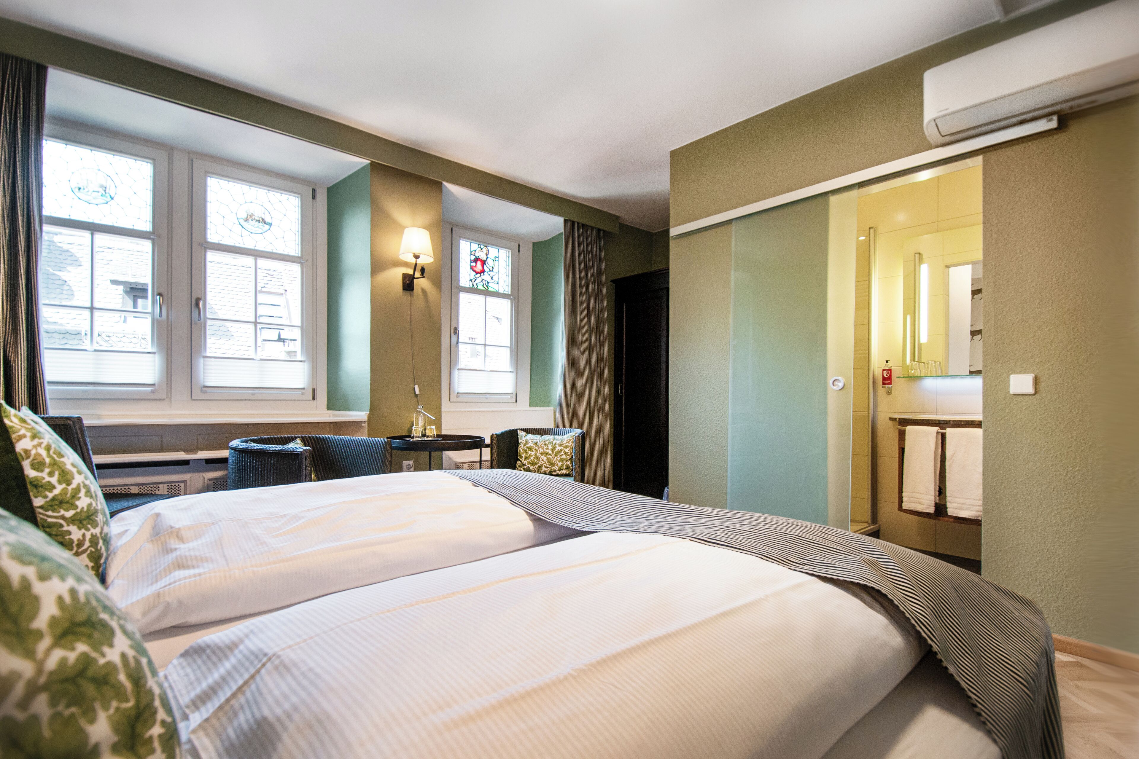 A hotel room with two beds, a desk, a chair, a window with curtains, a bathroom with a towel rack, and a wall-mounted air conditioner.