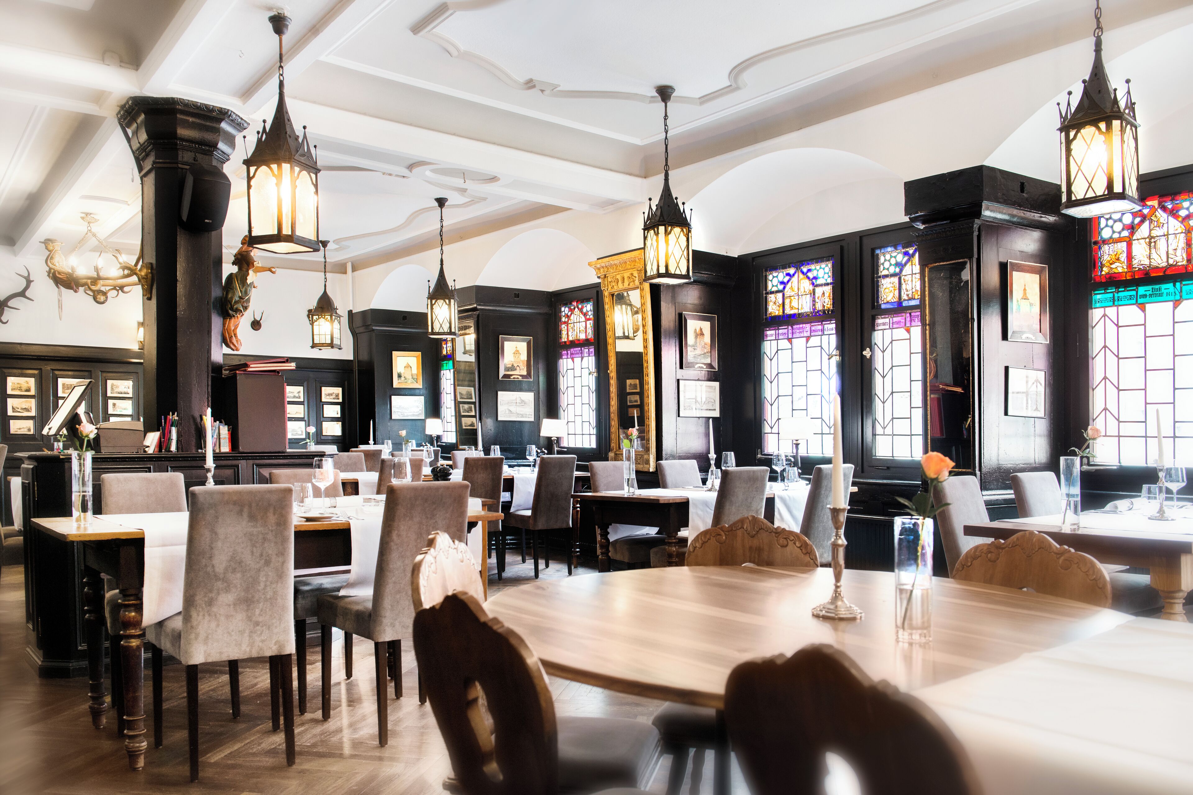 A restaurant with multiple dining tables and chairs, ornate chandeliers, stained glass windows, and framed pictures on the walls.