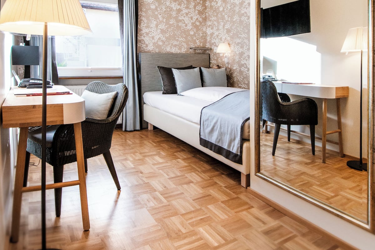 A modern hotel room with a bed, a desk, a chair, a large mirror, and a lamp.