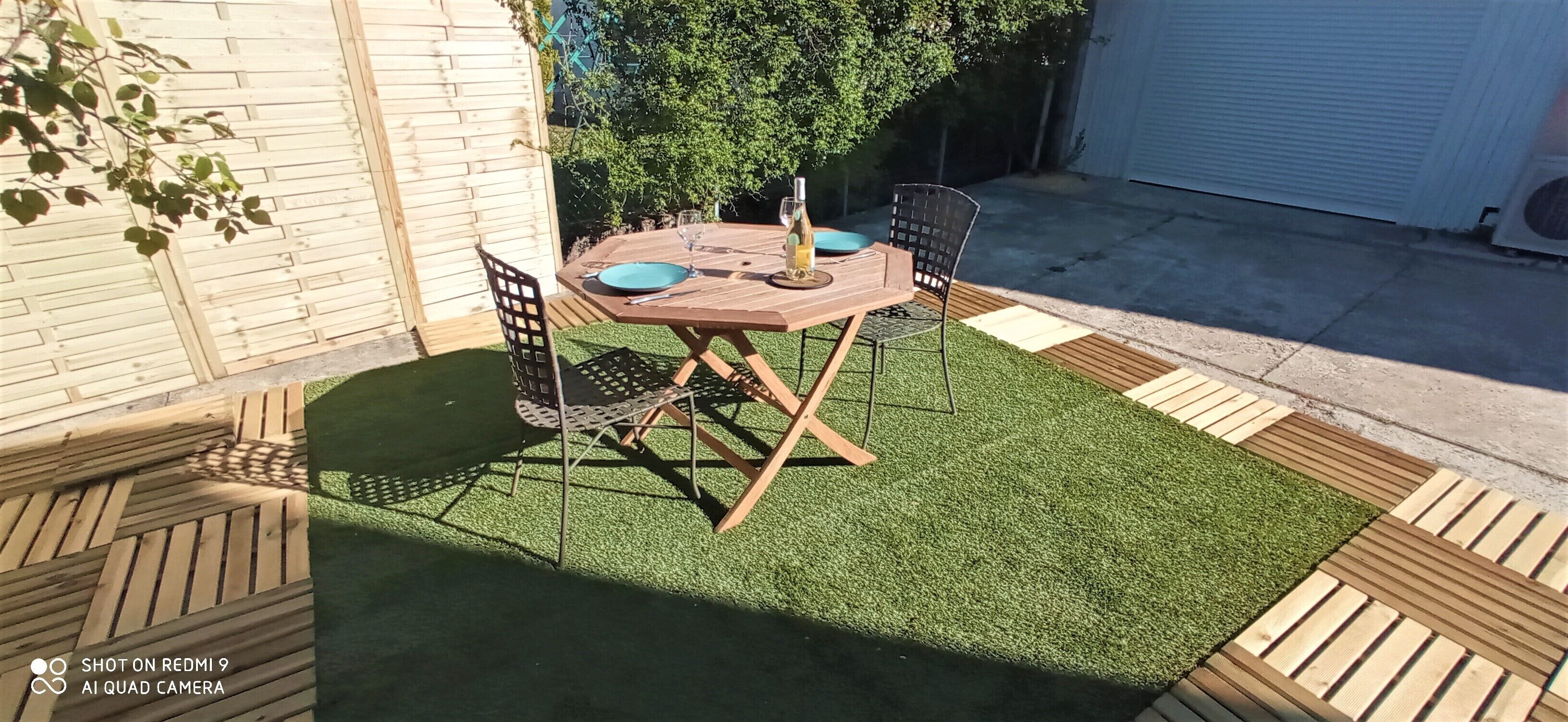 An outdoor patio with a wooden table and chairs, surrounded by artificial grass and wooden borders.