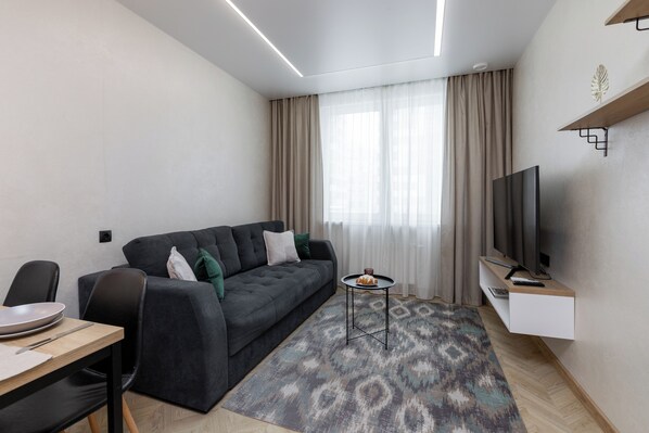 Een moderne woonkamer met een donkergrijze bank, een klein rond tafeltje, een flatscreen televisie en een kamerkleed met een patroon.