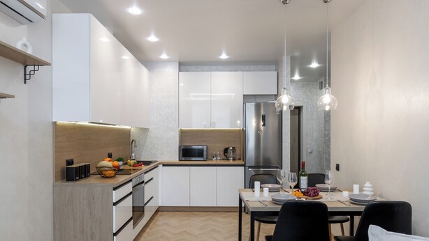 Una cocina moderna con armarios blancos, un microondas y un refrigerador de acero inoxidable. Hay un comedor con una mesa lista para dos personas, con un cuenco de fruta y copas de vino.