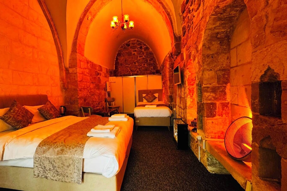 A hotel room with a stone wall, a bed, a second bed, a fireplace, and a chandelier.