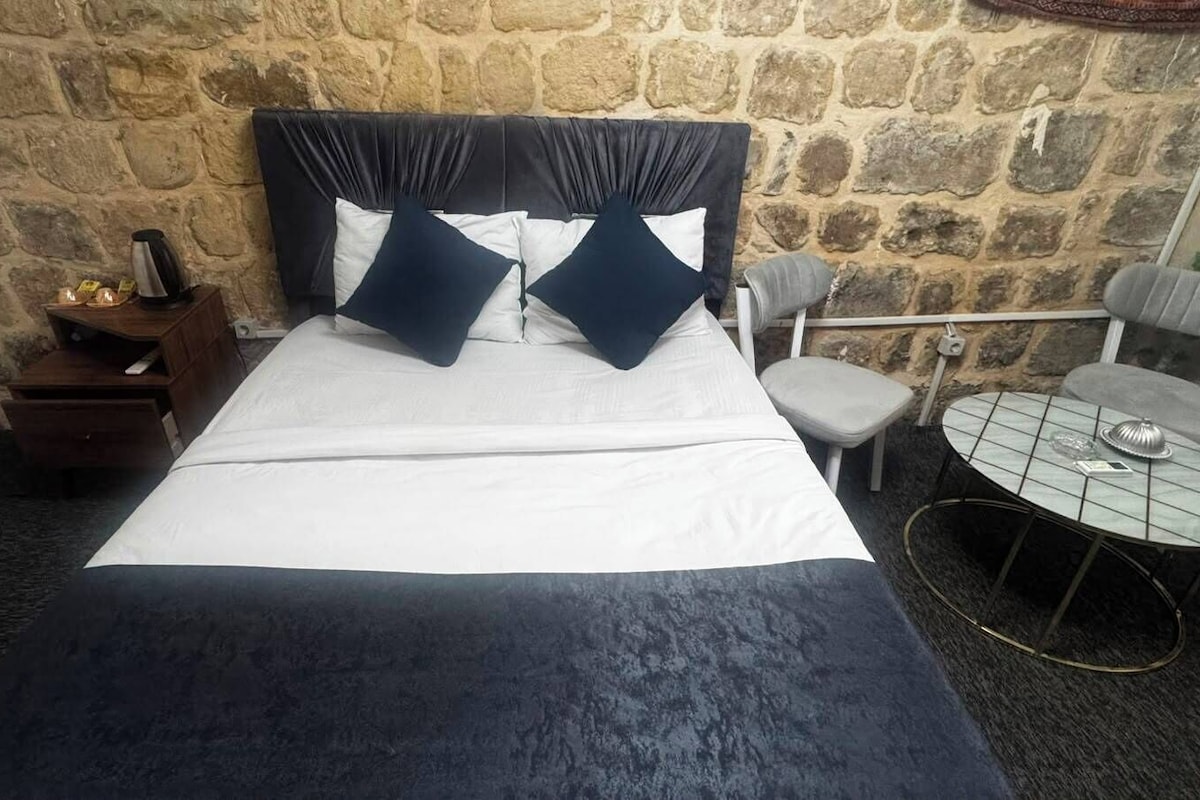 A bed with a dark headboard, white bedding, and two dark pillows. There is a small wooden nightstand with a kettle and a lamp on it. A round table with a metal frame and a chair is visible.