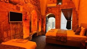 A room with stone walls, a bed with an orange bedspread, a flat-screen TV mounted on the wall, and a window with curtains. - Tunç Paşa Konağı (Mardin)