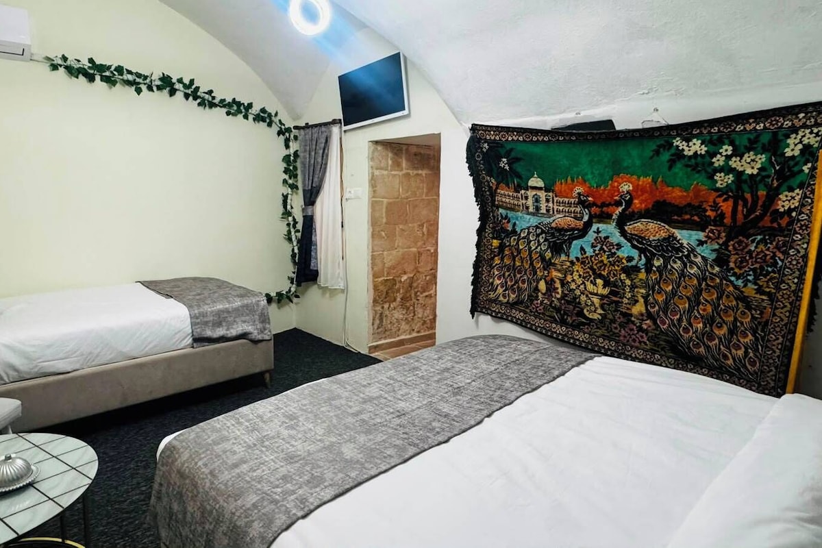 A room with two beds, a small table, a chair, a TV mounted on the wall, and a tapestry with a peacock design.