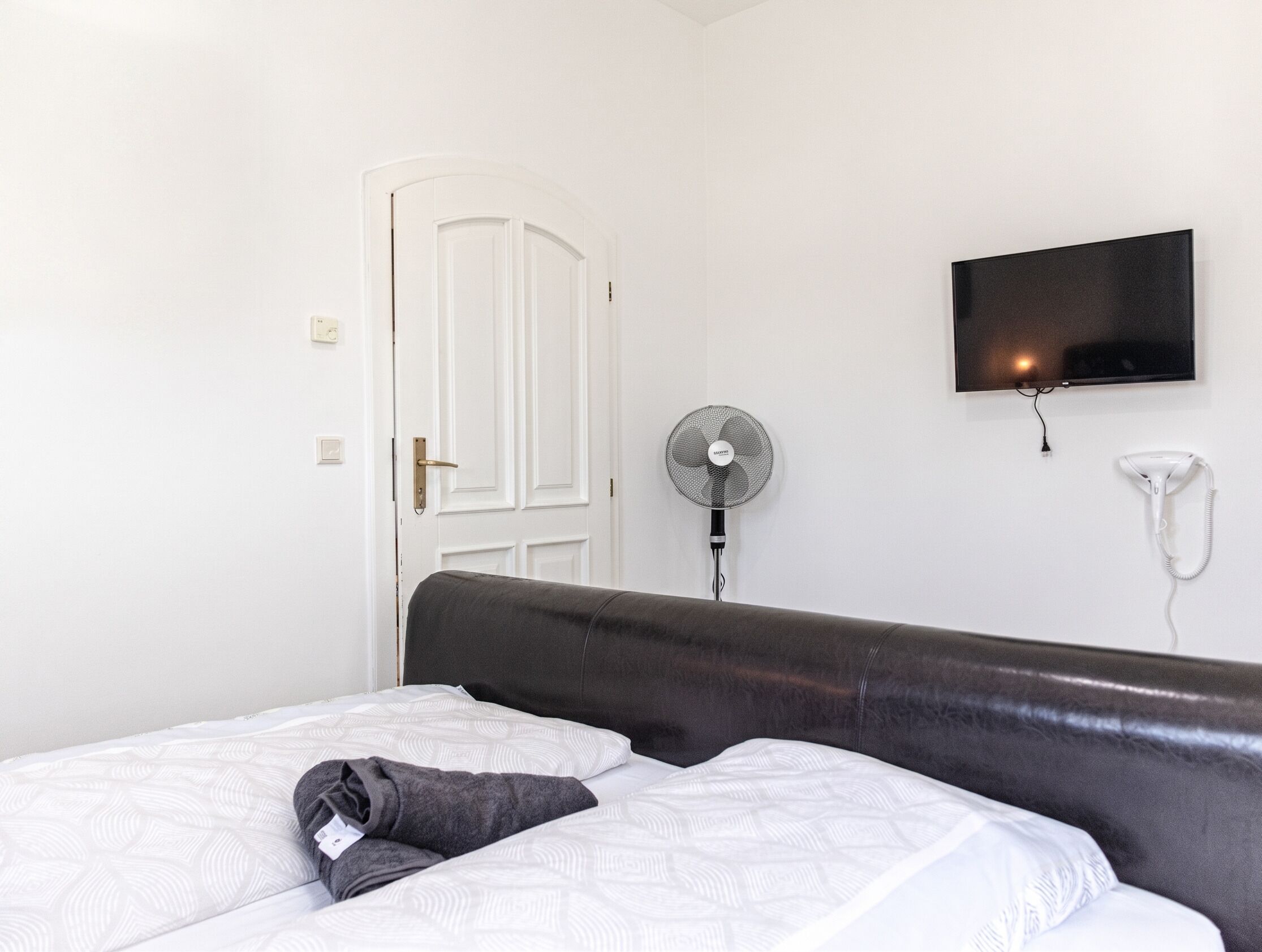 Un dormitorio con una cama, televisión montada en la pared, ventilador de pie y secador de pelo.