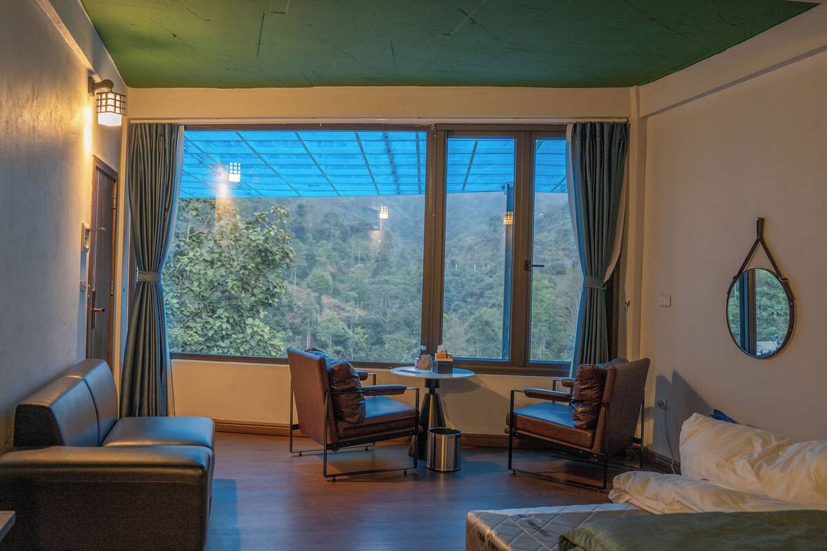 A room with a sofa, two armchairs, a small table, and a bed. There is a large window with curtains and a view of a forested area.
