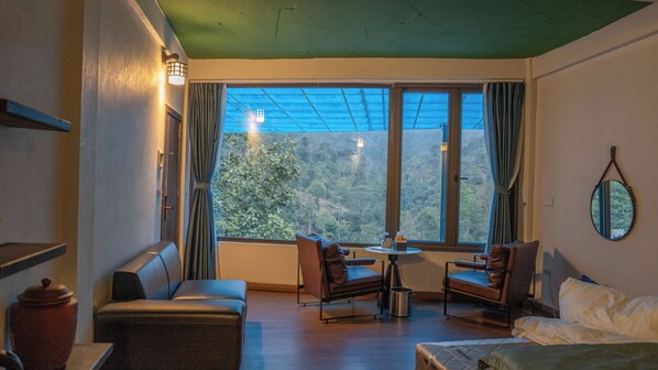 Une pièce comprenant un canapé, deux fauteuils, une petite table et un lit. Une grande fenêtre, ornée de rideaux, offre une vue sur une zone boisée.