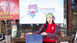 A person standing behind a counter with a blue gift bag, a glass display case, and a sign with a dragon logo. - The Legend of Dragon Tea Suoi Giang  (Van Chan)