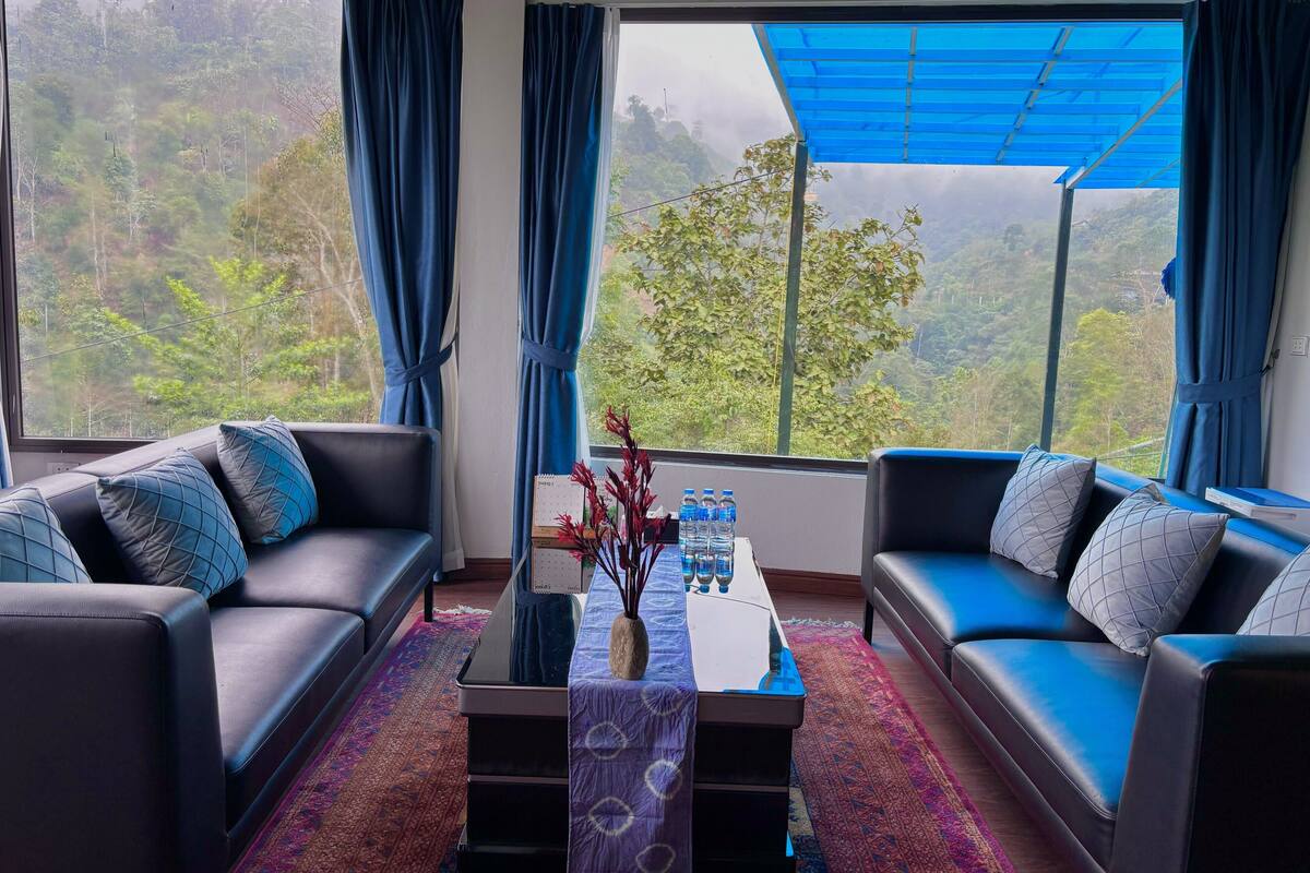 A modern living room with two black sofas, a glass coffee table, and a view of a lush green landscape through large windows.