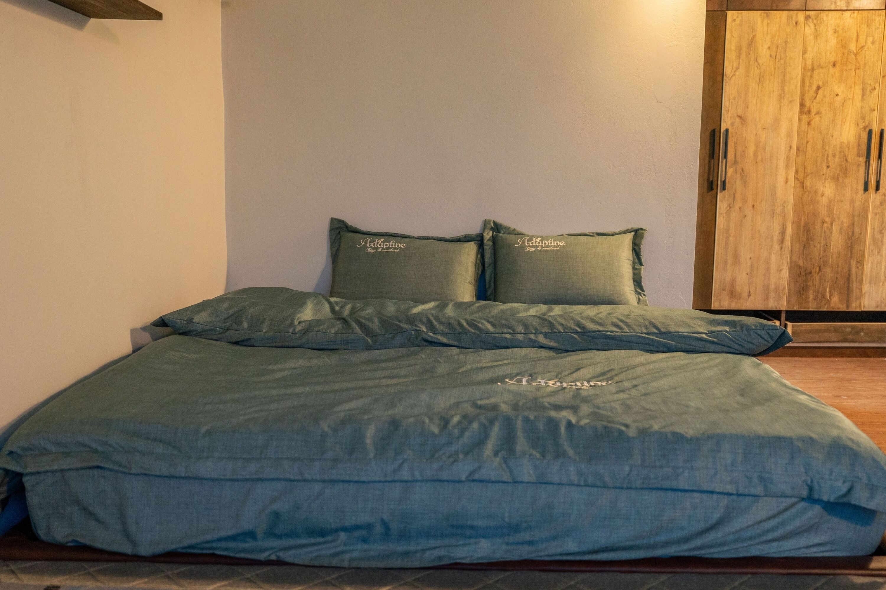 Un lit avec une couvre-lit verte et des oreillers, une armoire en bois et un sol carrelé.