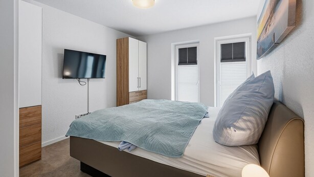 Ein Schlafzimmer mit einem Bett, einem an der Wand montierten Fernseher und einem Fenster mit Jalousien.
