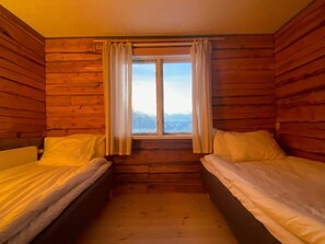 A cabin room with two beds, wooden walls, a window with curtains, and a view of the outdoors.