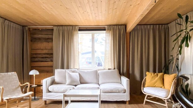 Ein Wohnzimmer mit einem weißen Sofa, einem Holztisch, einem weißen Sessel und einem gelben Sessel. Ein Fenster mit Vorhängen, eine Lampe und eine Pflanze sind ebenfalls vorhanden.