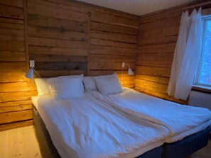 Ein holzgetäfeltes Zimmer im Blockhausstil mit einem Bett, zwei Nachttischlampen und einem Fenster mit Vorhängen.