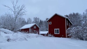 Ein rotes Holzhaus mit einem mit Schnee bedeckten Dach, umgeben von schneebedeckten Bäumen und Sträuchern.