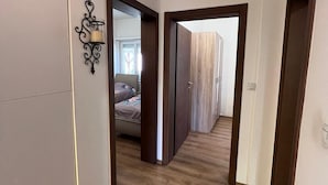 A hallway with wooden flooring, a bed in the background, and a wall-mounted light fixture. - Spacious 3-Bedroom, 2-Bath Apartment in Ilidža – Near Airport, Garden & BBQ (Ilidža)