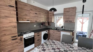 A modern kitchen with wooden cabinets, a gas stove, a washing machine, and a dining area with a patterned tablecloth. - Spacious 3-Bedroom, 2-Bath Apartment in Ilidža – Near Airport, Garden & BBQ (Ilidža)