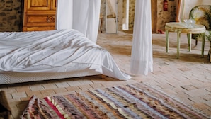 Une chambre à coucher rustique avec un mur en pierre, une porte en bois, un lit à baldaquin et une petite table ronde agrémentée d’une plante.